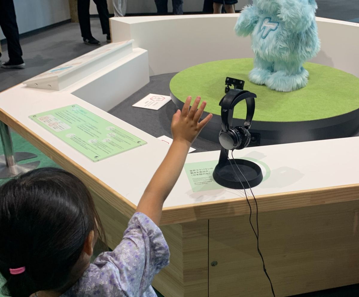 A child reaching toward a fluffy blue robot on a green turntable exhibit at Miraikan